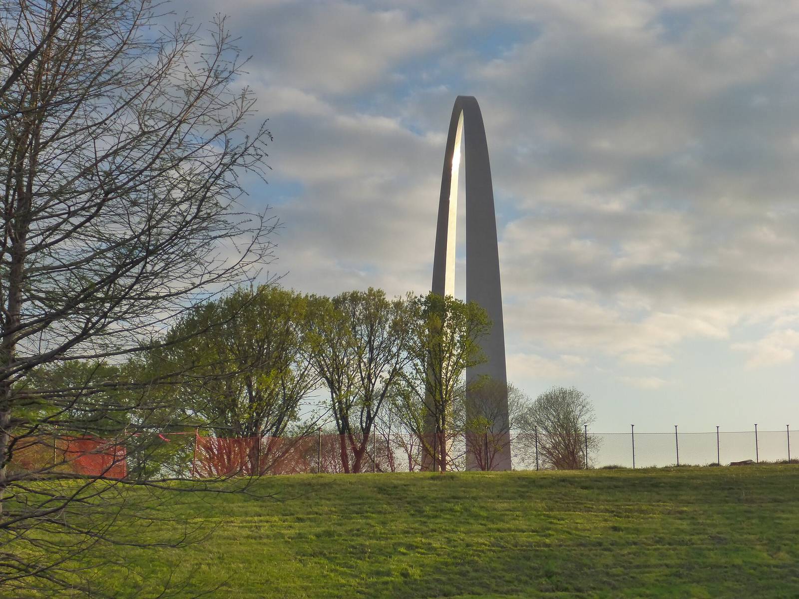 The Gateway Arch at sunrise viewed across the grounds, trees in spring leaf, a grass slope in the foreground.